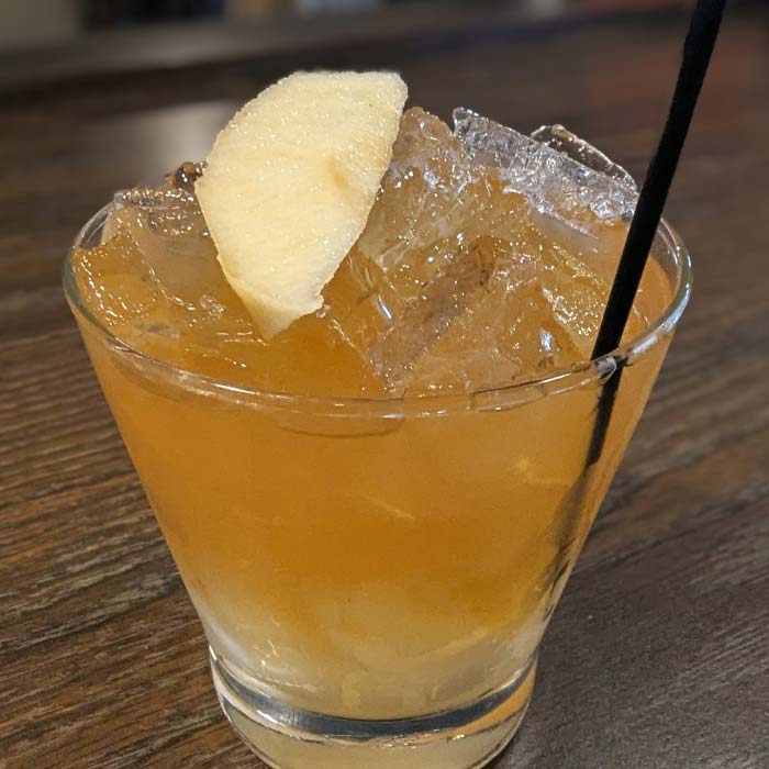 A cocktail with ice and apple slice at River’s Edge Supper Club, served on a wooden table.